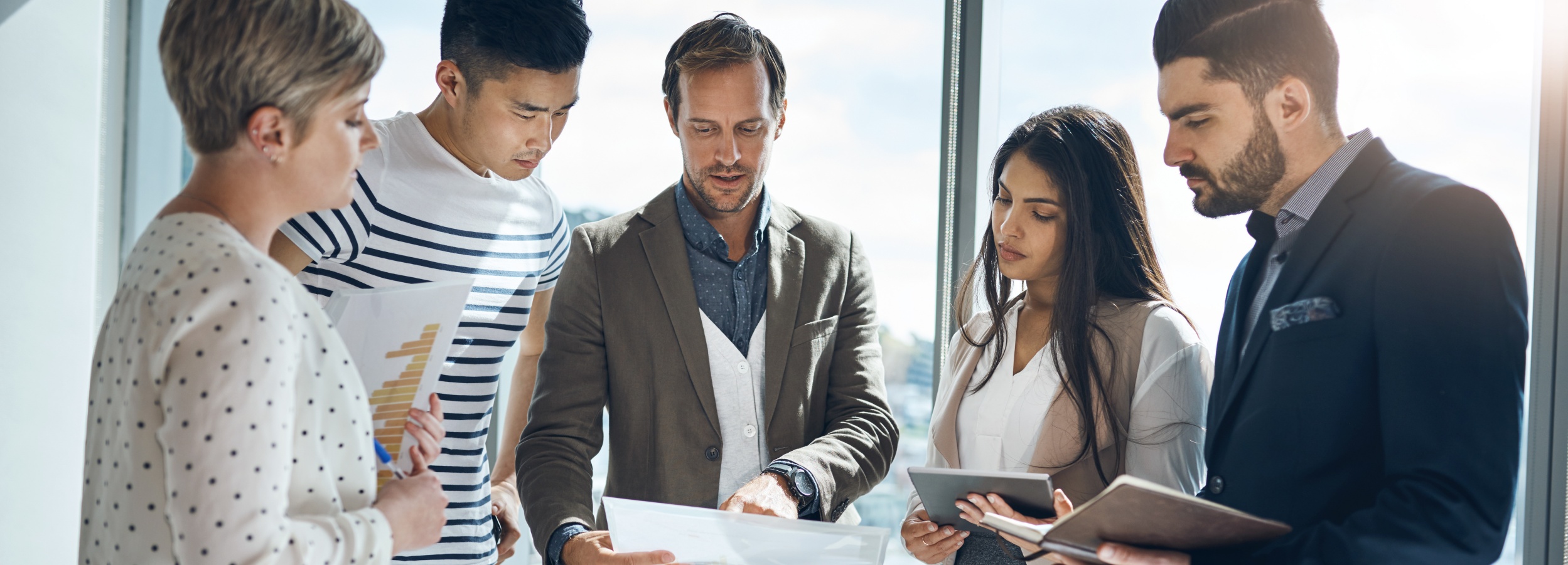 Eine Gruppe von fünf Fachleuten steht in einem hellen, modernen Büro zusammen und konzentriert sich auf Dokumente und Geräte in ihren Händen.

Das Bild zeigt fünf Personen, die in einem Büro stehen, wobei das Licht durch ein großes Fenster im Hintergrund einfällt. Von links nach rechts sind zu sehen: eine Frau mit kurzem, blondem Haar, die ein weißes Hemd mit schwarzen Punkten trägt, ein junger Mann mit einem gestreiften T-Shirt, ein Mann in einem grauen Sakko über einem hellblauen Hemd und einer weißen Weste, eine Frau mit langen, dunklen Haaren, die ein weißes Oberteil und eine beige Jacke trägt und ein Tablet in der Hand hält, und ein Mann mit Bart in einem dunklen Anzug. Die Personen scheinen in eine Diskussion oder Analyse von Dokumenten verwickelt zu sein, da sie alle nach unten auf die Dokumente und Geräte schauen. Die Atmosphäre ist professionell und konzentriert, mit einem Hauch von collaborativem Teamwork.