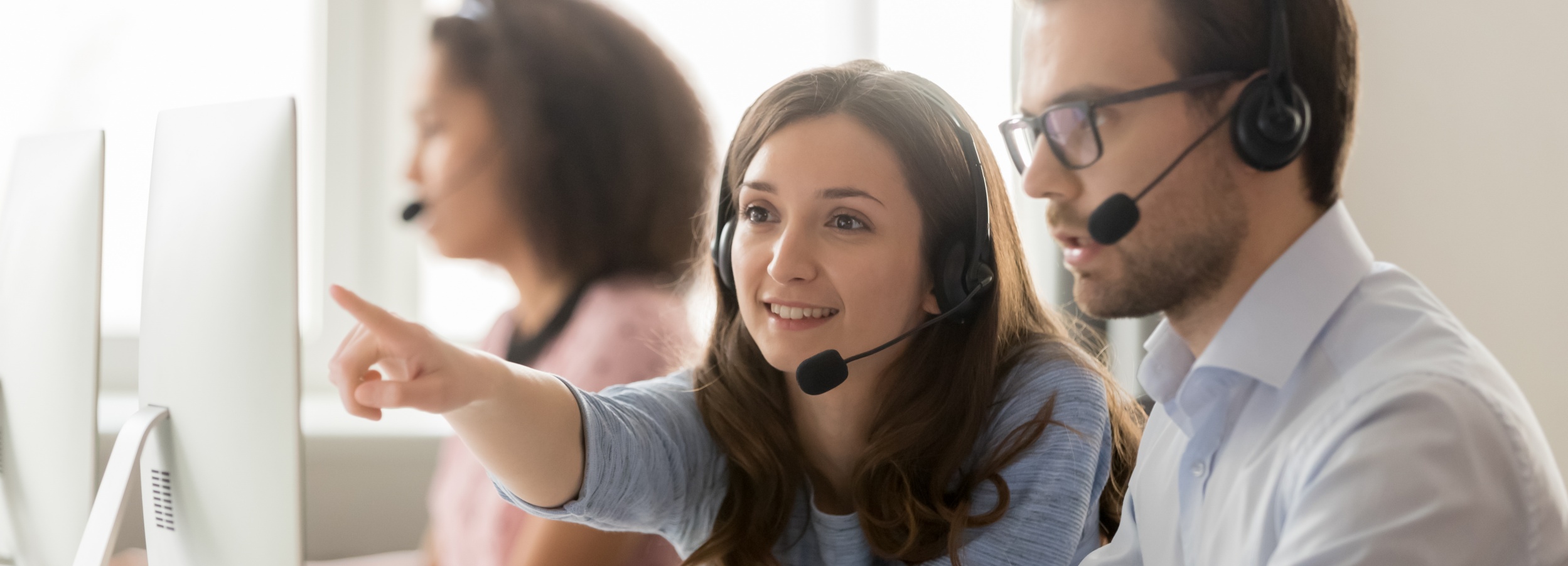 Das Bild zeigt eine Gruppe von Kundendienstmitarbeitern bei der Arbeit, wobei sich ein Mann und eine Frau in den Vordergrund rücken, während sie auf einen Computerbildschirm schauen und Anweisungen geben.

Das Bild zeigt drei Kundendienstmitarbeiter, die alle Headsets tragen und vor Computern sitzen. Im Vordergrund sind eine Frau und ein Mann zu sehen. Die Frau zeigt mit dem Finger auf den Computerbildschirm und lächelt, während der Mann aufmerksam zuhört. Beide tragen formelle Kleidung: Die Frau ein hellblaues Hemd und der Mann ein weißes Hemd mit Brille. Im Hintergrund ist eine weitere Mitarbeiterin zu sehen, ebenfalls mit Headset, aber unscharf. Die Atmosphäre wirkt professionell und kooperativ, mit einem positiven und hilfsbereiten Unterton. Die Beleuchtung ist hell und gleichmäßig, was die Gesichter der Mitarbeiter gut sichtbar macht. Die Farbtöne sind sanft und beruhigend, was eine angenehme Arbeitsumgebung suggeriert.