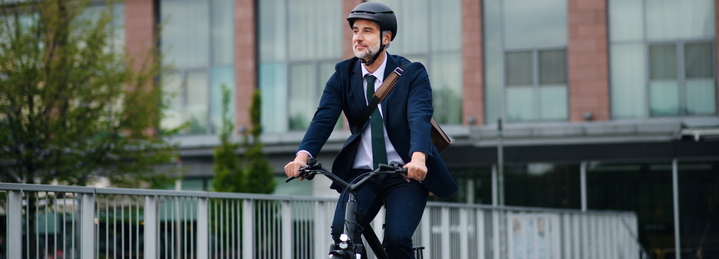 Ein grauhaariger Mann in Anzug und Helm fährt mit einem Fahrrad durch die Stadt.

Das Bild zeigt einen erwachsenen Mann mittleren Alters, der einen dunklen Anzug, ein weißes Hemd und eine grüne Krawatte trägt und auf einem Fahrrad sitzt. Er hat graue Haare und einen Bart und trägt einen schwarzen Helm. Seine Hände halten den Lenker des Fahrrads fest, und er blickt lächelnd nach rechts. Auf seiner rechten Schulter trägt er eine braune Umhängetasche. Das Fahrrad selbst ist dunkel und scheint ein moderneres Modell zu sein, mit einem Scheinwerfer vorne. Der Mann befindet sich auf einer Brücke oder einem erhöhten Weg, der von einem metallenen Geländer gesäumt wird. Im Hintergrund ist ein großes Gebäude mit roten Ziegelsteinfassaden und großflächigen Fenstern zu sehen. Vor dem Gebäude stehen Bäume mit hellem, grünem Laub. Die Atmosphäre des Bildes ist optimistisch und dynamisch, was durch den Blick des Mannes und seine Kleidung, die eine Mischung aus professioneller Eleganz und aktiver Mobilität darstellt, unterstrichen wird. Die Beleuchtung scheint diffus zu sein, was auf einen bewölkten Tag oder eine spätere Tagesstunde hindeutet, aber die Szene wirkt dennoch lebendig.