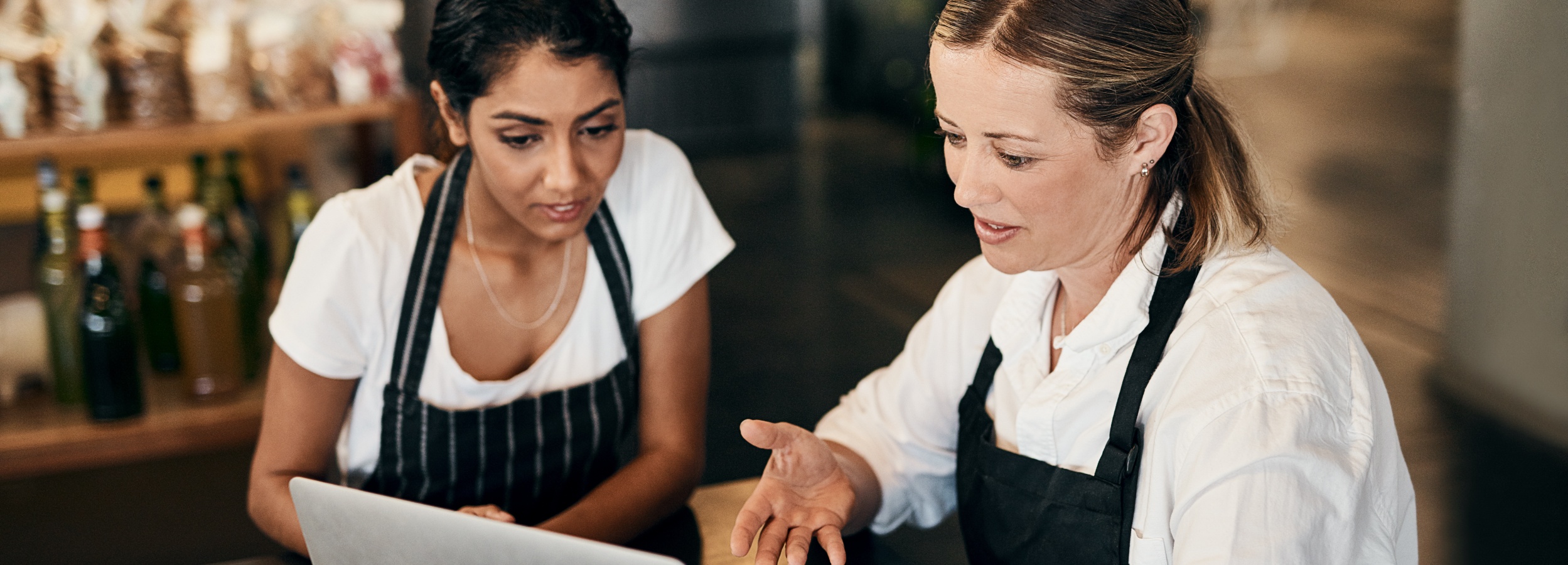 Zwei Frauen, offenbar Angestellte eines Geschäfts, besprechen etwas anhand eines Laptops in einem beleuchteten, aber gedämpften Umfeld.

Auf dem Bild sind zwei Frauen zu sehen, die sich in einem Geschäft befinden, das wahrscheinlich in der Lebensmittel- oder Getränkebranche tätig ist, da im Hintergrund Regale mit Flaschen und Gläsern zu erkennen sind. Beide Frauen tragen eine weiße Kleidung und eine schwarze Schürze, was auf ihre Tätigkeit in diesem Geschäft hindeutet. Die Frau auf der linken Seite, mit dunklen, kurz geschnittenen Haaren und dunkler Haut, blickt konzentriert auf einen Laptop, der vor ihnen auf einer Oberfläche steht. Ihre Augen sind gesenkt, und ihr Gesichtsausdruck wirkt nachdenklich. Sie trägt eine schlichte silberne Halskette. Die Frau auf der rechten Seite, mit helleren Haaren, die zu einem niedrigen Pferdeschwanz zurückgebunden sind, hat den Blick ebenfalls auf den Laptop gerichtet und gestikuliert mit der rechten Hand, als würde sie etwas erklären oder einen Punkt hervorheben. Ihr Gesichtsausdruck ist engagiert und gesprächig. Sie trägt ebenfalls eine weiße Bluse und eine schwarze Schürze sowie einen kleinen Ohrstecker und eine filigrane Halskette. Die Beleuchtung im Vordergrund ist warm und fokussiert auf die beiden Frauen und den Laptop, während der Hintergrund in weicherem Licht liegt und die Objekte auf den Regalen nur schemenhaft erkennbar sind, was eine intime und fokussierte Atmosphäre schafft. Die Stimmung des Bildes ist professionell und kooperativ, was auf eine gemeinsame Arbeit und Problemlösung oder Planung hindeutet.