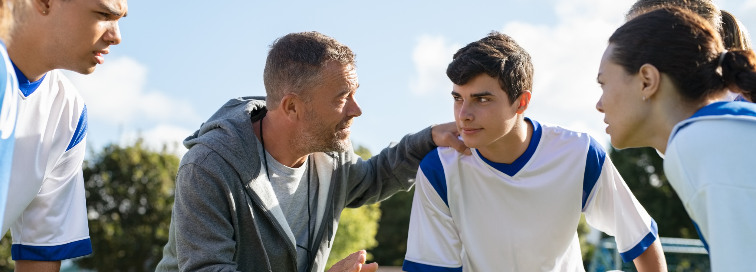 Ein Fußballtrainer gibt seinem Team Anweisungen vor dem Spiel oder in der Halbzeitpause.

Das Bild zeigt einen Fußballtrainer, der mit einigen seiner Spieler spricht. Der Fokus liegt auf dem Trainer, einem Mann mittleren Alters mit grauen Haaren, der eine graue Kapuzenjacke über einem grauen T-Shirt trägt. Er legt seine Hand auf die Schulter eines jungen Spielers, der ihm direkt zuhört. Der Spieler trägt ein weißes Fußballtrikot mit blauen Akzenten an den Schultern und am Kragen.

Um die beiden herum sind weitere Spieler zu sehen, die teilweise angeschnitten sind. Man erkennt auch, dass der Himmel leicht bewölkt ist und Bäume im Hintergrund stehen. Die Atmosphäre wirkt konzentriert und motivierend, da der Trainer seine Weisheit und Strategie mit dem Team teilt. Die Stimmung ist ernst, aber konstruktiv, da alle Beteiligten auf das gemeinsame Ziel fokussiert sind.