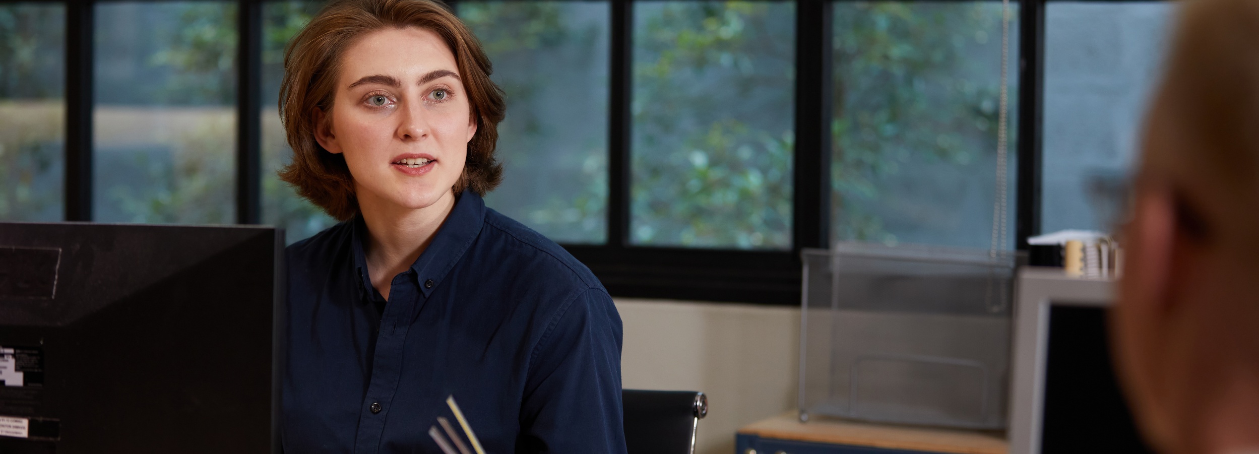 Das Bild zeigt eine junge Frau in einem Büro, die mit einem Kollegen spricht.

Im Detail zeigt das Bild den Oberkörper einer jungen Frau mit hellbraunen, schulterlangen Haaren, die ein dunkelblaues Hemd trägt. Sie sitzt an einem Schreibtisch, vor dem ein schwarzer Computermonitor steht. Ihr Blick ist nach rechts gewandt, wo man nur den Hinterkopf einer anderen blondhaarigen Person sieht, was darauf hindeutet, dass sie sich in einem Gespräch befindet. Im Hintergrund ist ein Fenster zu sehen, das einen Blick ins Grüne freigibt, was dem Raum eine beruhigende Atmosphäre verleiht. Auf der rechten Seite des Bildes sind Teile eines weiteren Schreibtisches mit Bürobedarf zu erkennen. Die Atmosphäre wirkt professionell und gleichzeitig entspannt, was durch das natürliche Licht und die Pflanzen im Hintergrund verstärkt wird.