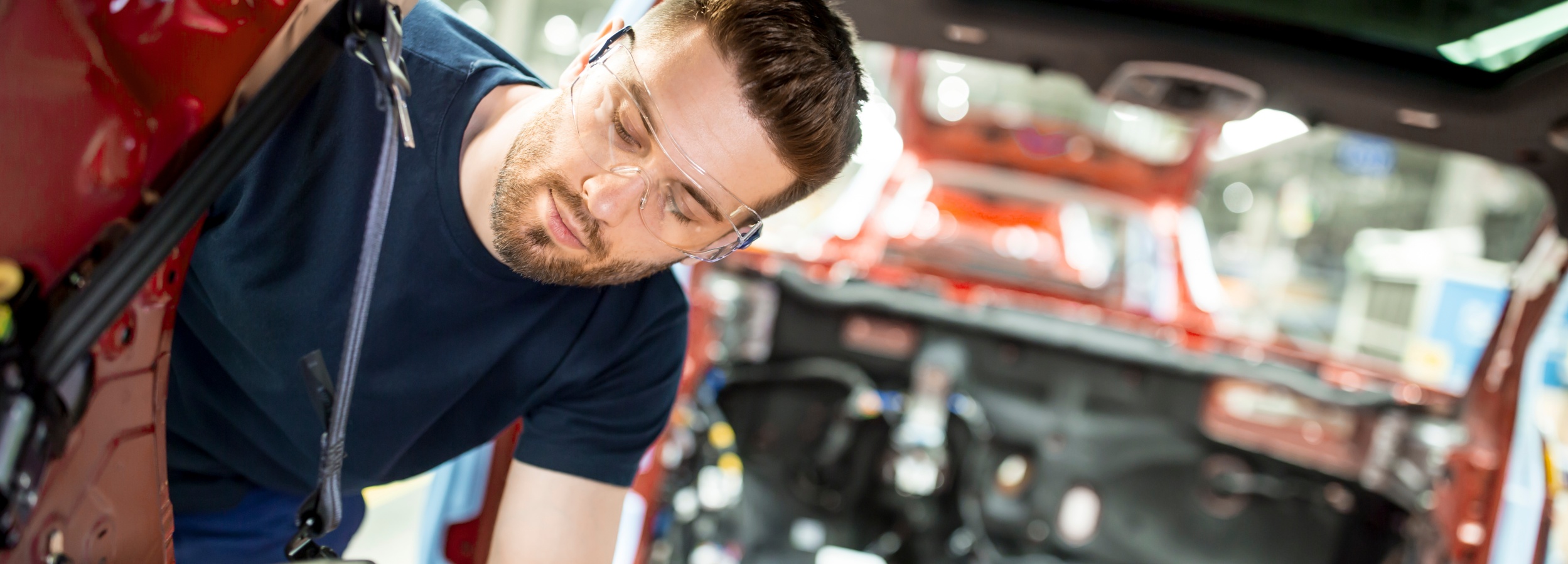 Ein Mann mit Schutzbrille arbeitet konzentriert an der Innenausstattung eines roten Autos. 

Das Bild zeigt einen Mann mit Bart und kurzen braunen Haaren, der eine Schutzbrille trägt und in ein rotes Auto hineinlehnt. Er scheint an der Inneneinrichtung oder Verkabelung des Fahrzeugs zu arbeiten. Sein Gesicht ist nach unten gerichtet, und seine Augen sind auf die Arbeit konzentriert. Er trägt ein dunkelblaues T-Shirt.

Der Hintergrund zeigt das Innere des Autos, das in leuchtendem Rot lackiert ist, sowie verschiedene Kabel und mechanische Teile, die zugänglich sind. Das Sonnenlicht scheint von oben durch ein Schiebedach und beleuchtet den Innenraum. Die Atmosphäre ist hell und professionell, was auf eine Werkstatt oder Produktionsstätte hindeuten könnte.