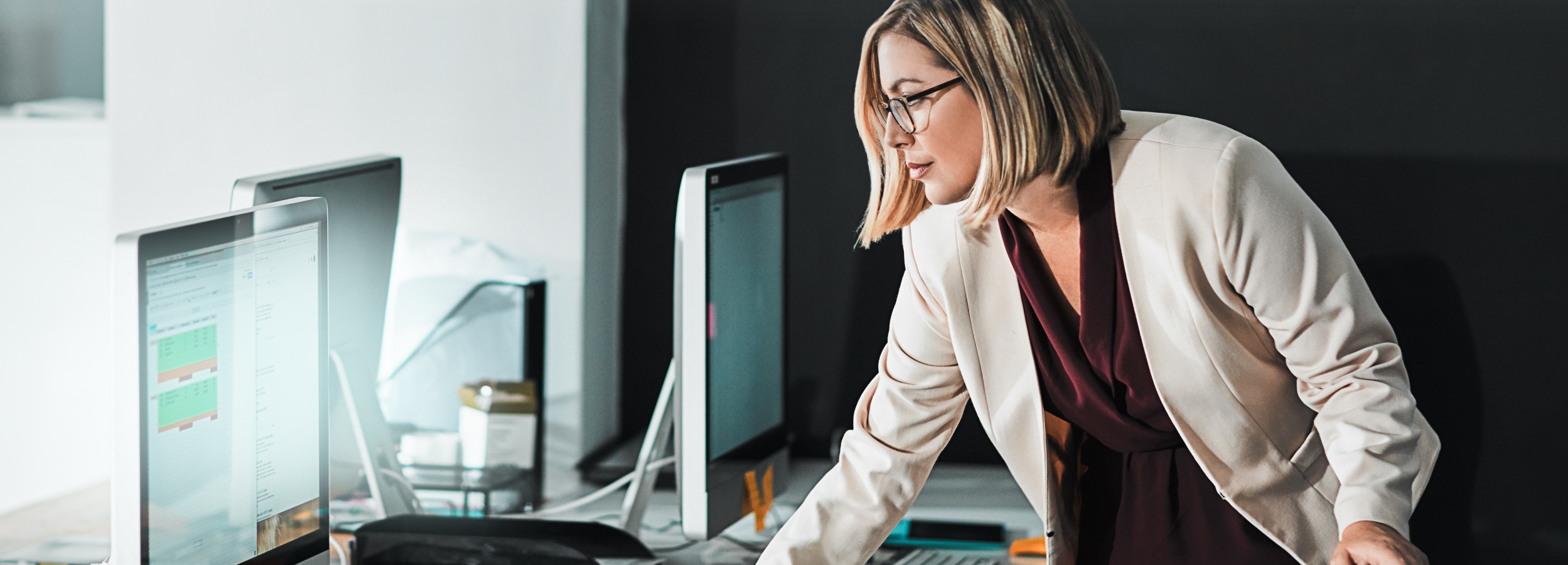 Eine professionell gekleidete Frau beugt sich über einen Schreibtisch voller Computerbildschirme und anderer Bürogegenstände, während sie konzentriert auf einen der Bildschirme schaut.

Das Bild zeigt eine blonde Frau mit Brille, die einen beigen Blazer und eine dunkelrote Bluse trägt. Sie steht an einem Schreibtisch, auf dem mehrere Computerbildschirme stehen. Auf den Bildschirmen sind verschiedene Fenster und Anwendungen zu sehen. Die Frau scheint sich auf den Inhalt eines der Bildschirme zu konzentrieren. Die Atmosphäre des Bildes ist professionell und konzentriert. Der Hintergrund ist unscharf und dunkel, was den Fokus auf die Frau und die Computerbildschirme lenkt. Das Bild vermittelt den Eindruck von harter Arbeit und Engagement im Büro.